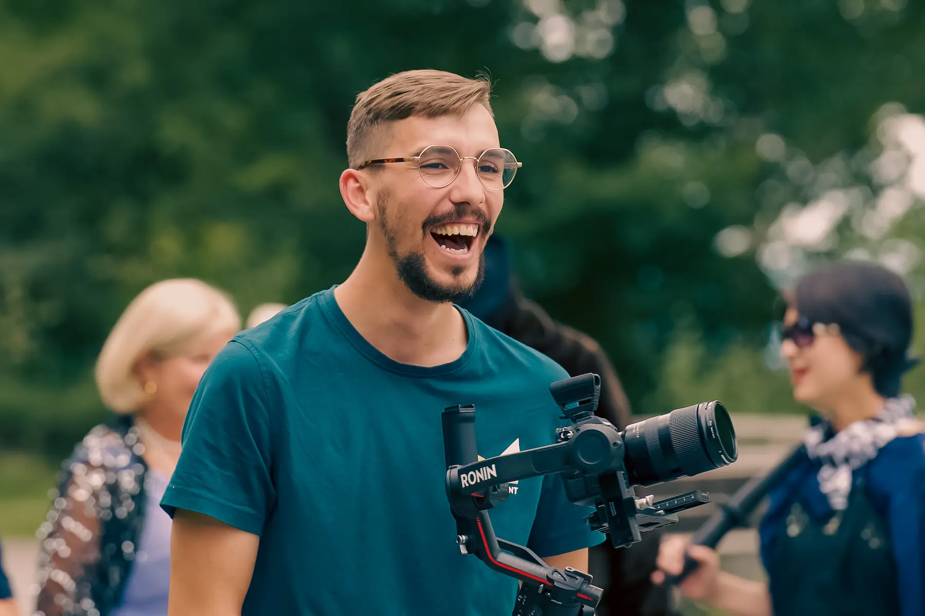 Portrait von einem jungen Mann mit einer professionellen Kamera in der Hand. Der Filmemacher von Filmevent lacht, im Hintergrund macht sich seine Gruppe bereit für die Nächste Szene.