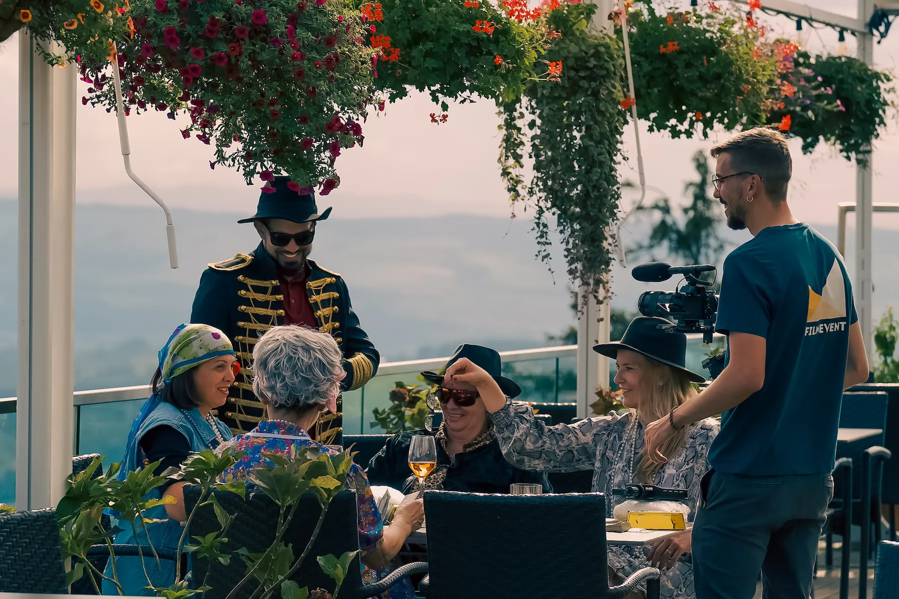 Eine Gruppe Menschen sitzt von Pflanzen umgeben und als Mafia Familie verkleidet an einem Tisch. Ein Filmemacher von Filmevent gibt der Gruppe Instruktionen. Die Leute lachen und machen sich bereit für die nächste Szene.